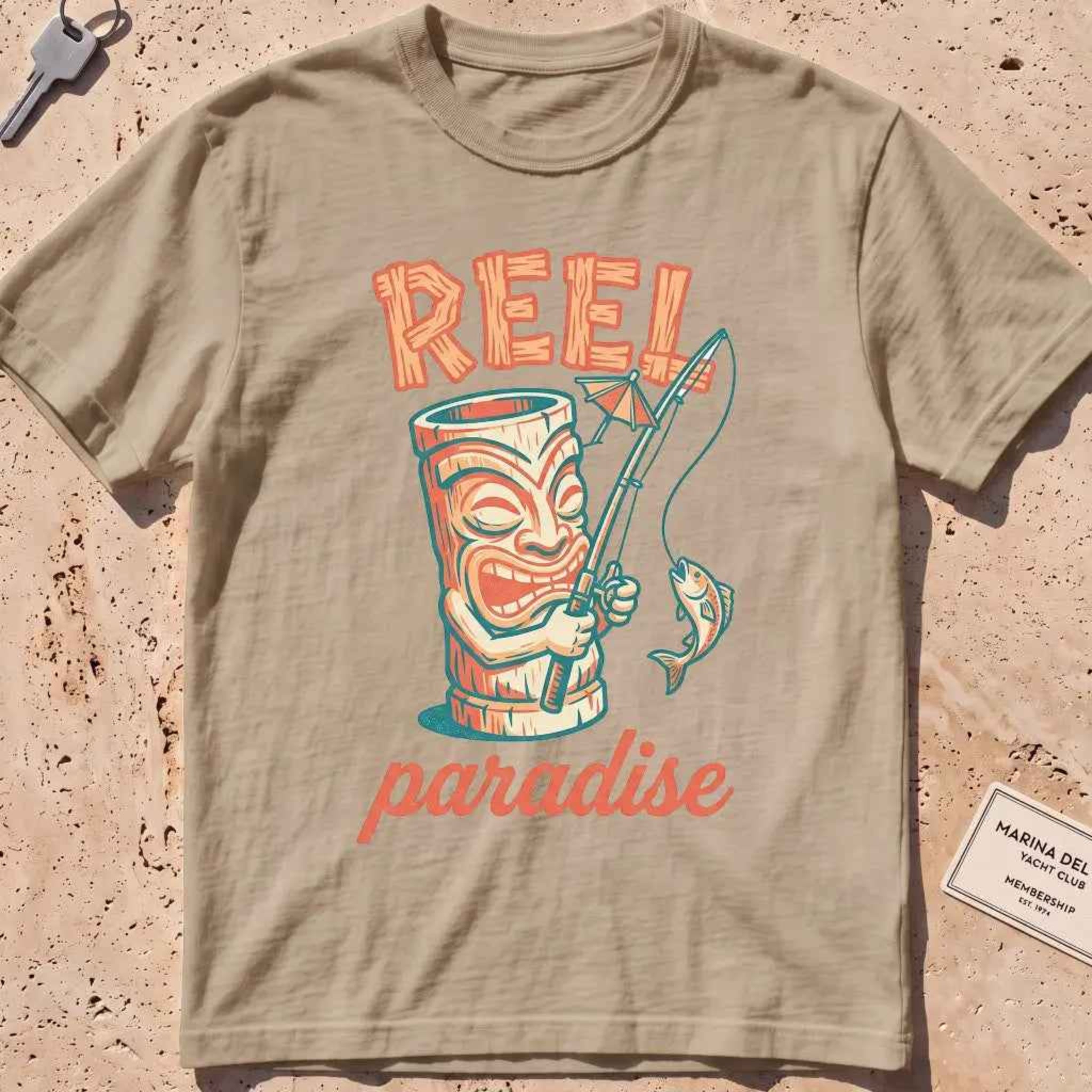 khaki heavyweight tee with a playful tiki mug and "Reel Paradise" text graphic.