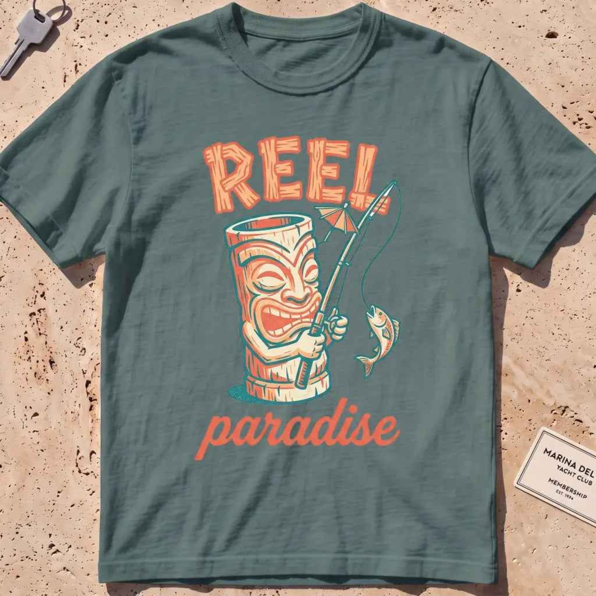blue spruce heavyweight tee with a playful tiki mug and "Reel Paradise" text graphic.