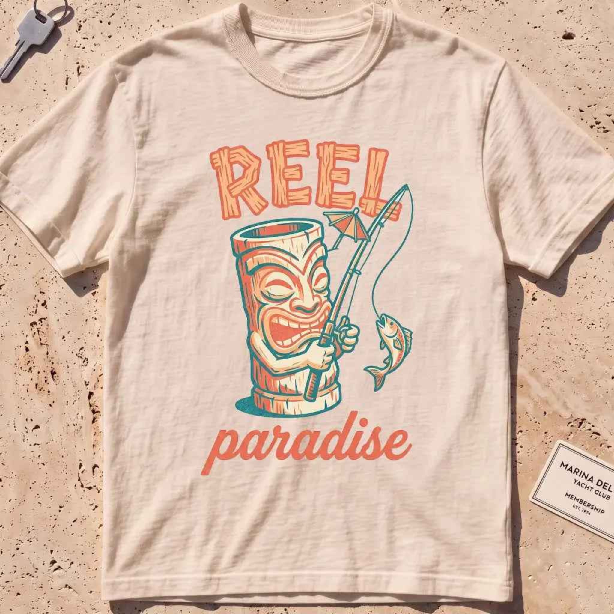 ivory heavyweight tee with a playful tiki mug and "Reel Paradise" text graphic.