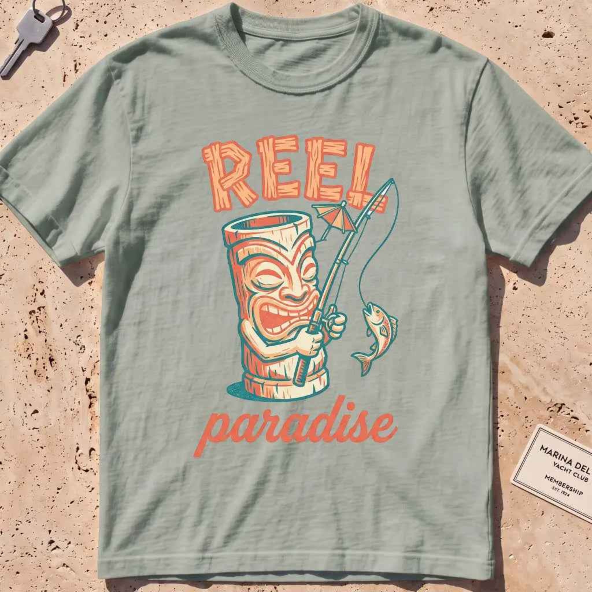 bay heavyweight tee with a playful tiki mug and "Reel Paradise" text graphic.