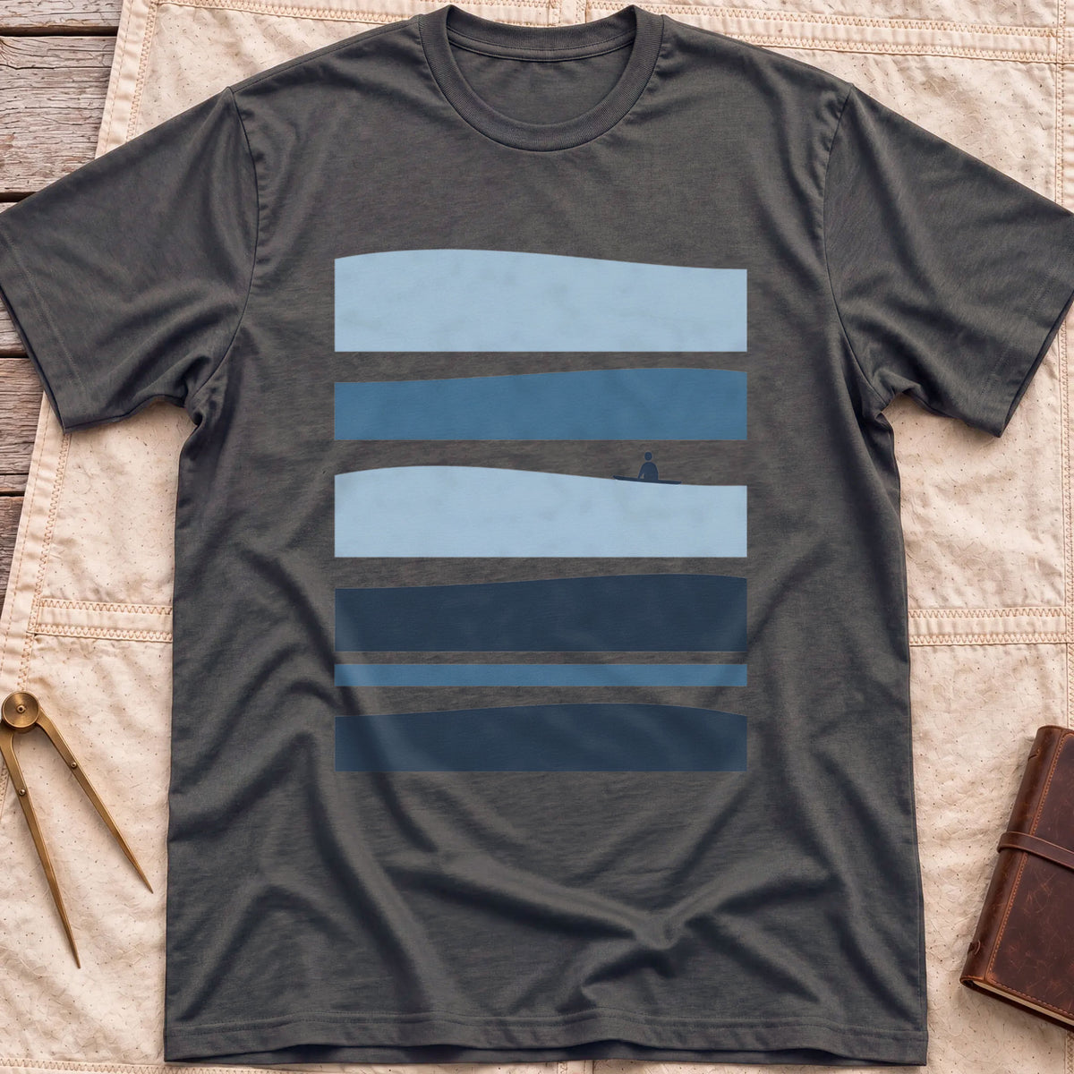 Front view of the dark heather Between Sets T-Shirt with layered bands of blue water and a small figure on a board between them.