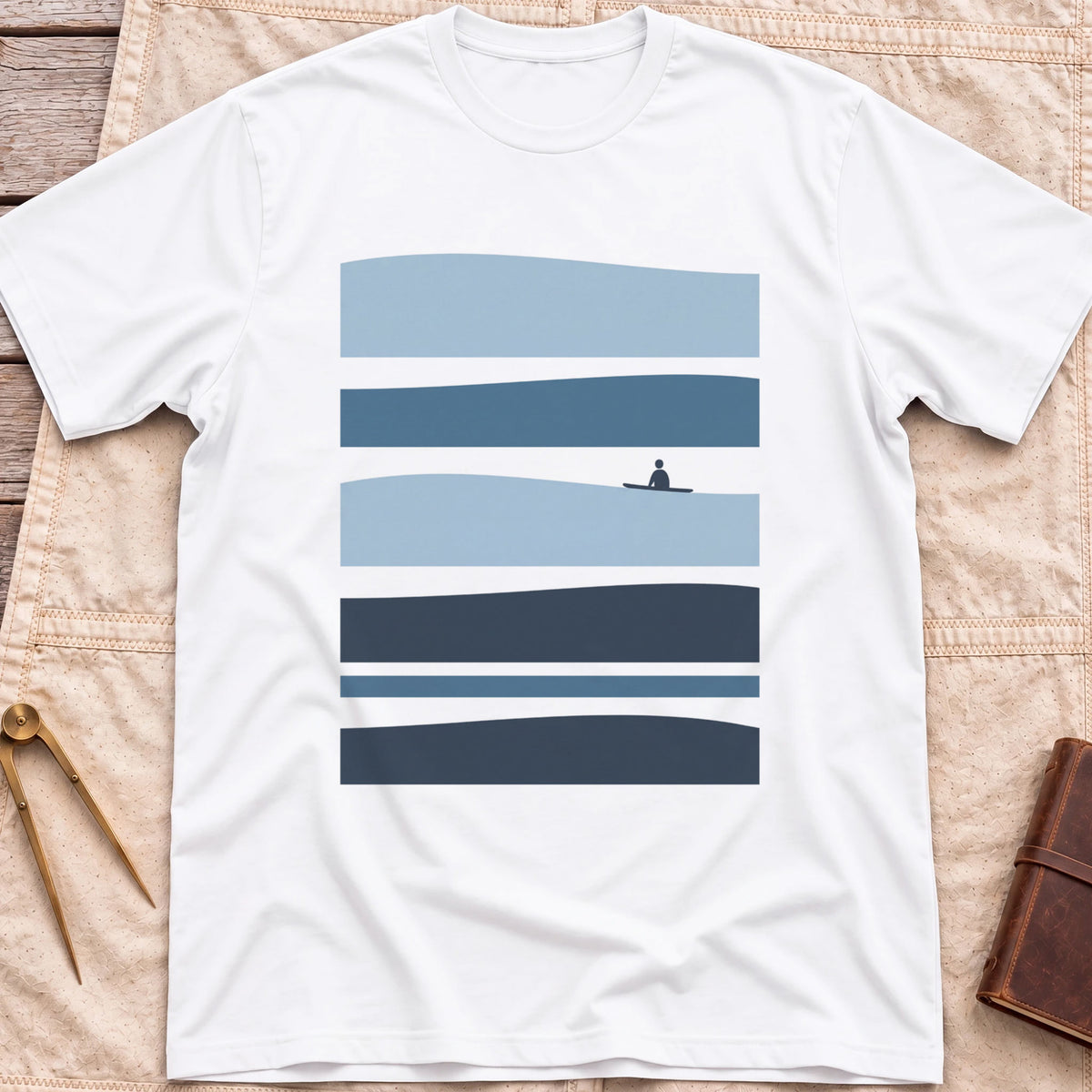 Front view of the white Between Sets T-Shirt with layered bands of blue water and a small figure on a board between them.