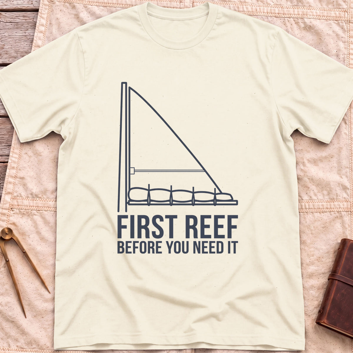 Front view of the natural First Reef T-Shirt with a line drawing of a sail with its lower section folded along the boom, above the words Before You Need It.