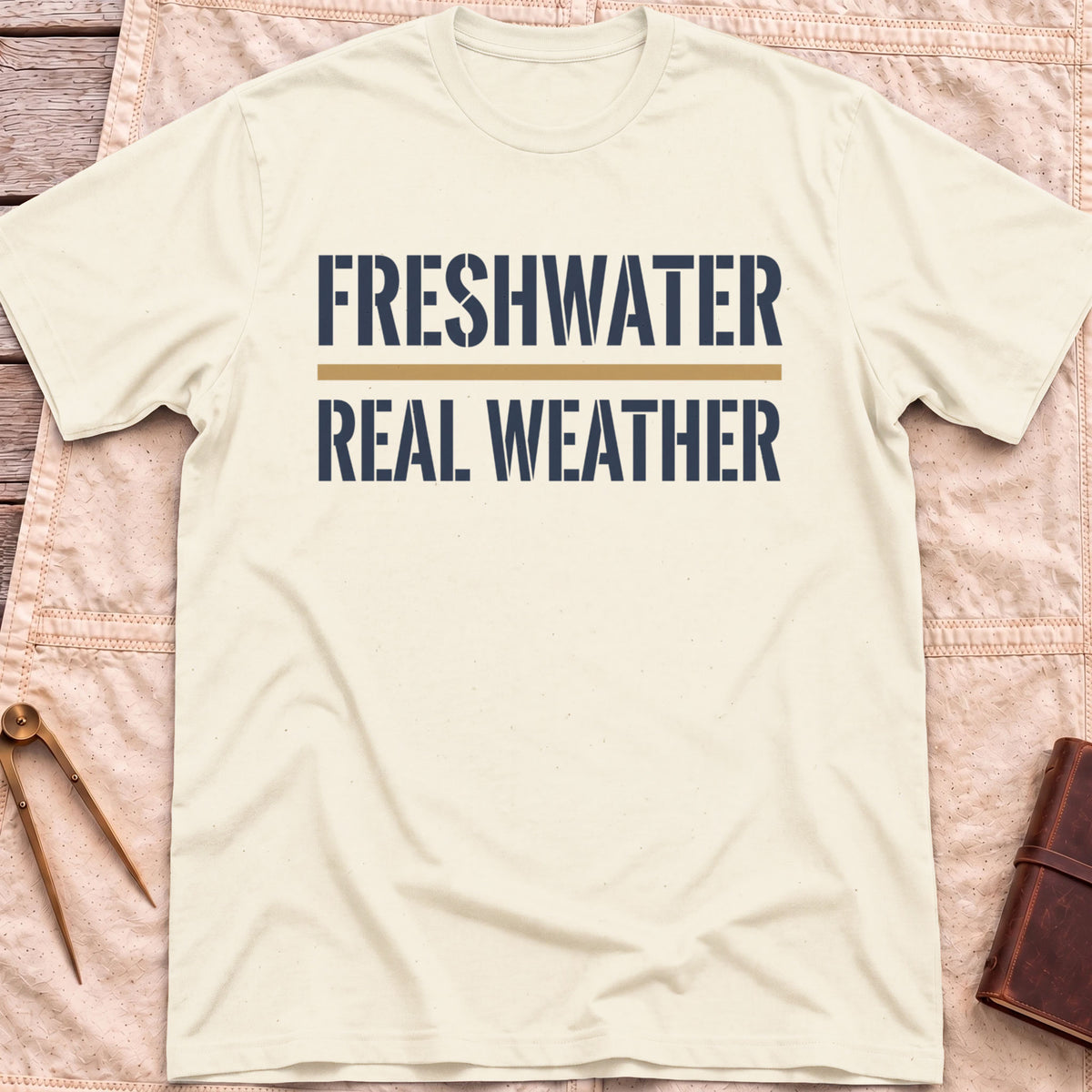 Front view of the natural Freshwater Real Weather T-Shirt with the words Freshwater and Real Weather in bold capitals, split by a tan line.