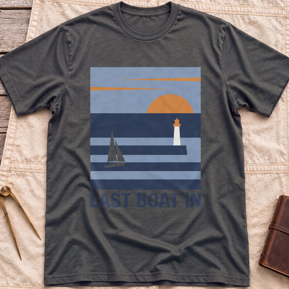 Front view of the dark heather Last Boat In T-Shirt with a square scene of a sailboat, a lighthouse and an orange sun over striped water.