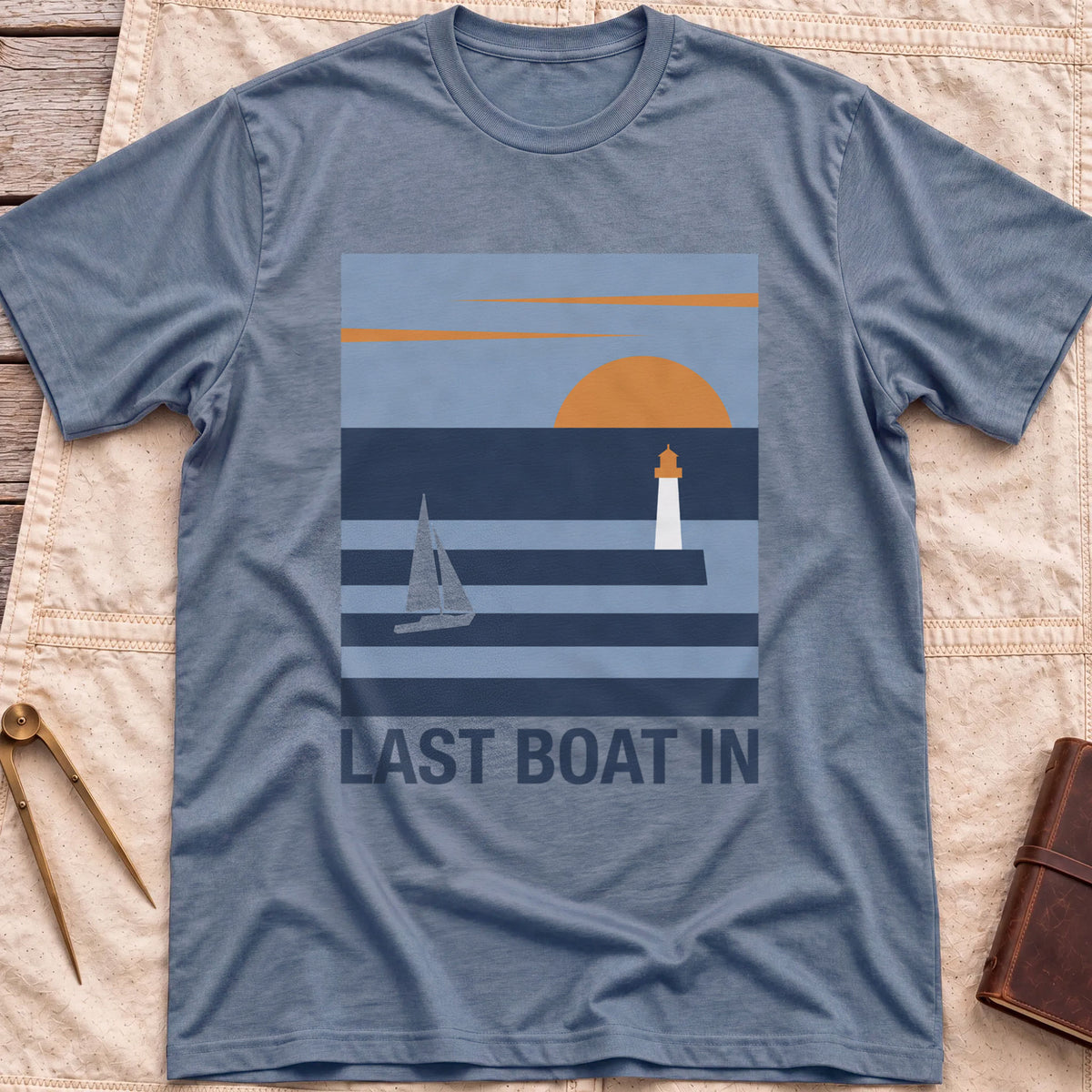 Front view of the heather indigo Last Boat In T-Shirt with a square scene of a sailboat, a lighthouse and an orange sun over striped water.