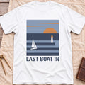 Front view of the white Last Boat In T-Shirt with a square scene of a sailboat, a lighthouse and an orange sun over striped water.