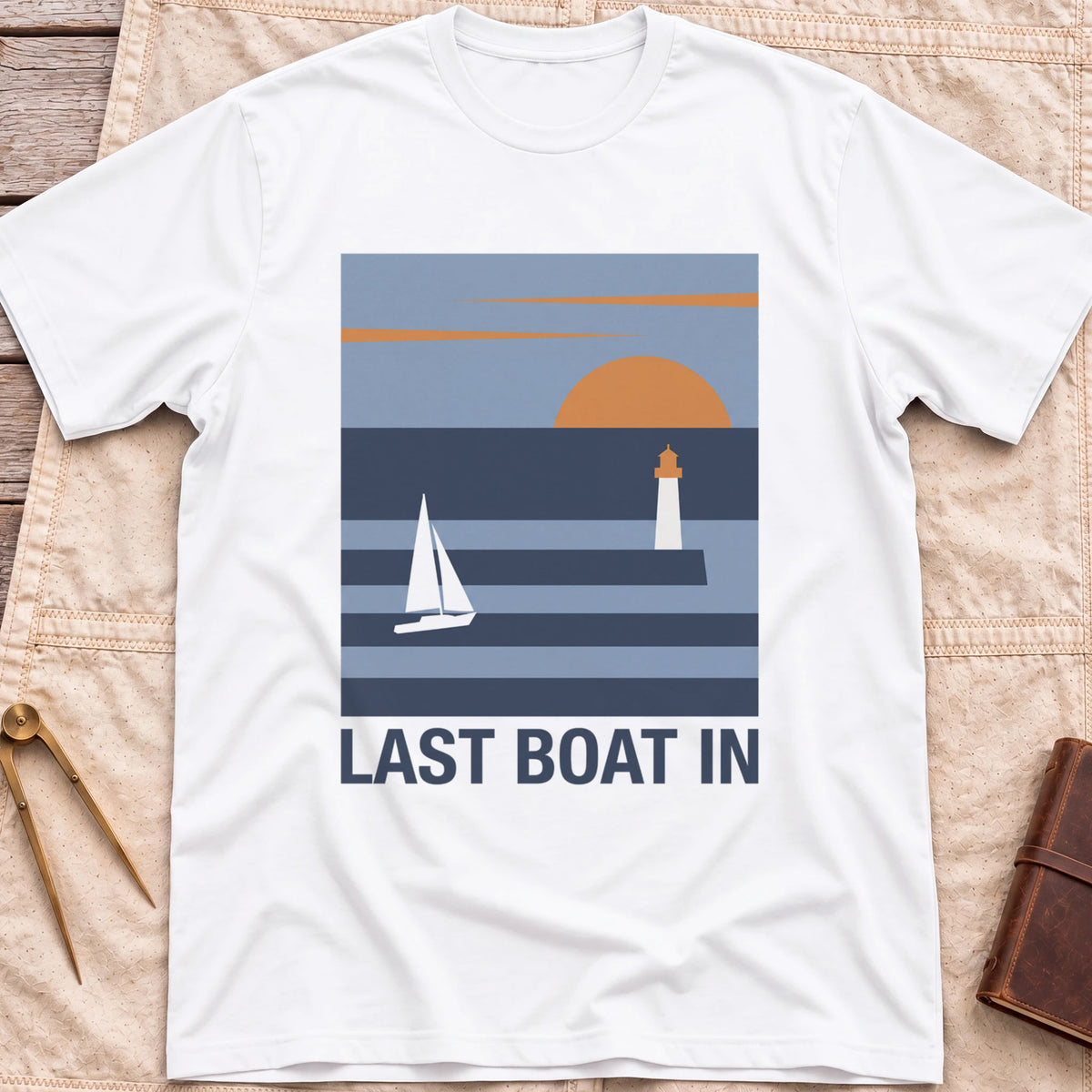 Front view of the white Last Boat In T-Shirt with a square scene of a sailboat, a lighthouse and an orange sun over striped water.