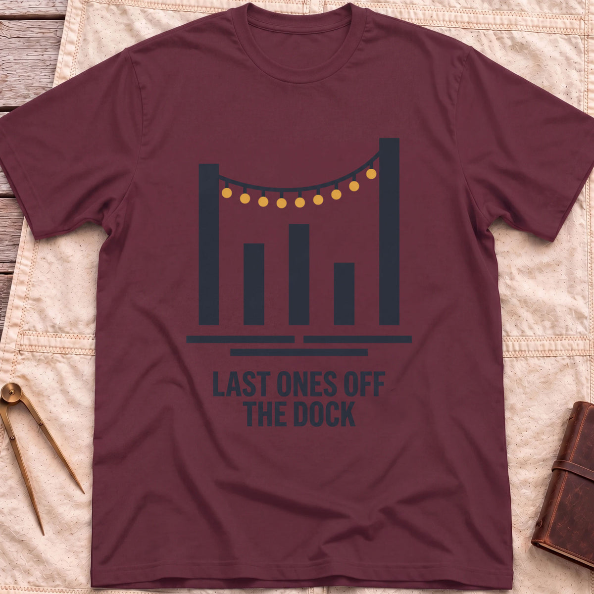 Front view of the maroon Last Ones Off the Dock T-Shirt with a string of lights hung between tall dock posts.