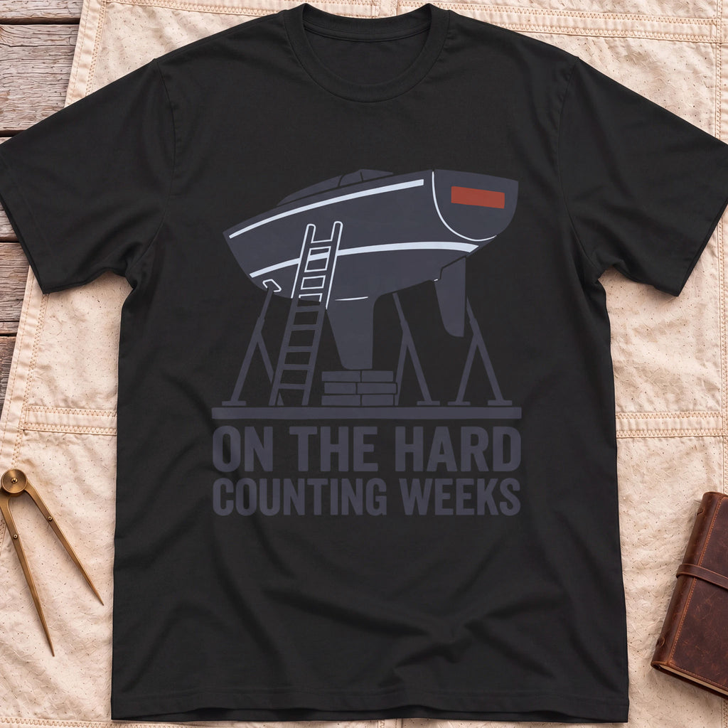 Front view of the black On the Hard T-Shirt with a boat hull up on stands with a ladder against it, above the words Counting Weeks.