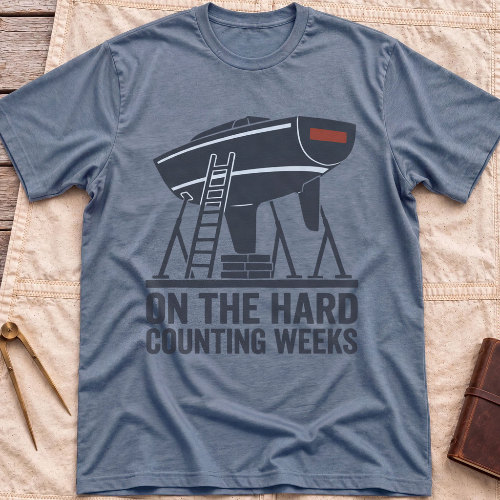 Front view of the heather indigo On the Hard T-Shirt with a boat hull up on stands with a ladder against it, above the words Counting Weeks.