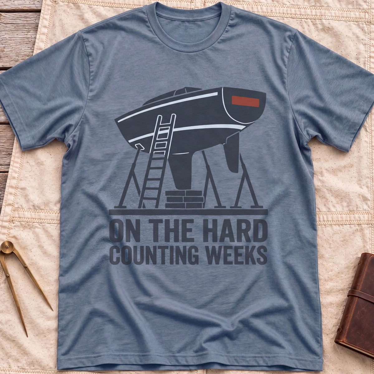 Front view of the heather indigo On the Hard T-Shirt with a boat hull up on stands with a ladder against it, above the words Counting Weeks.