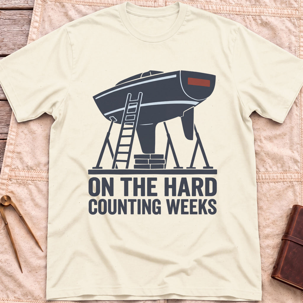 Front view of the natural On the Hard T-Shirt with a boat hull up on stands with a ladder against it, above the words Counting Weeks.