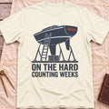Front view of the natural On the Hard T-Shirt with a boat hull up on stands with a ladder against it, above the words Counting Weeks.