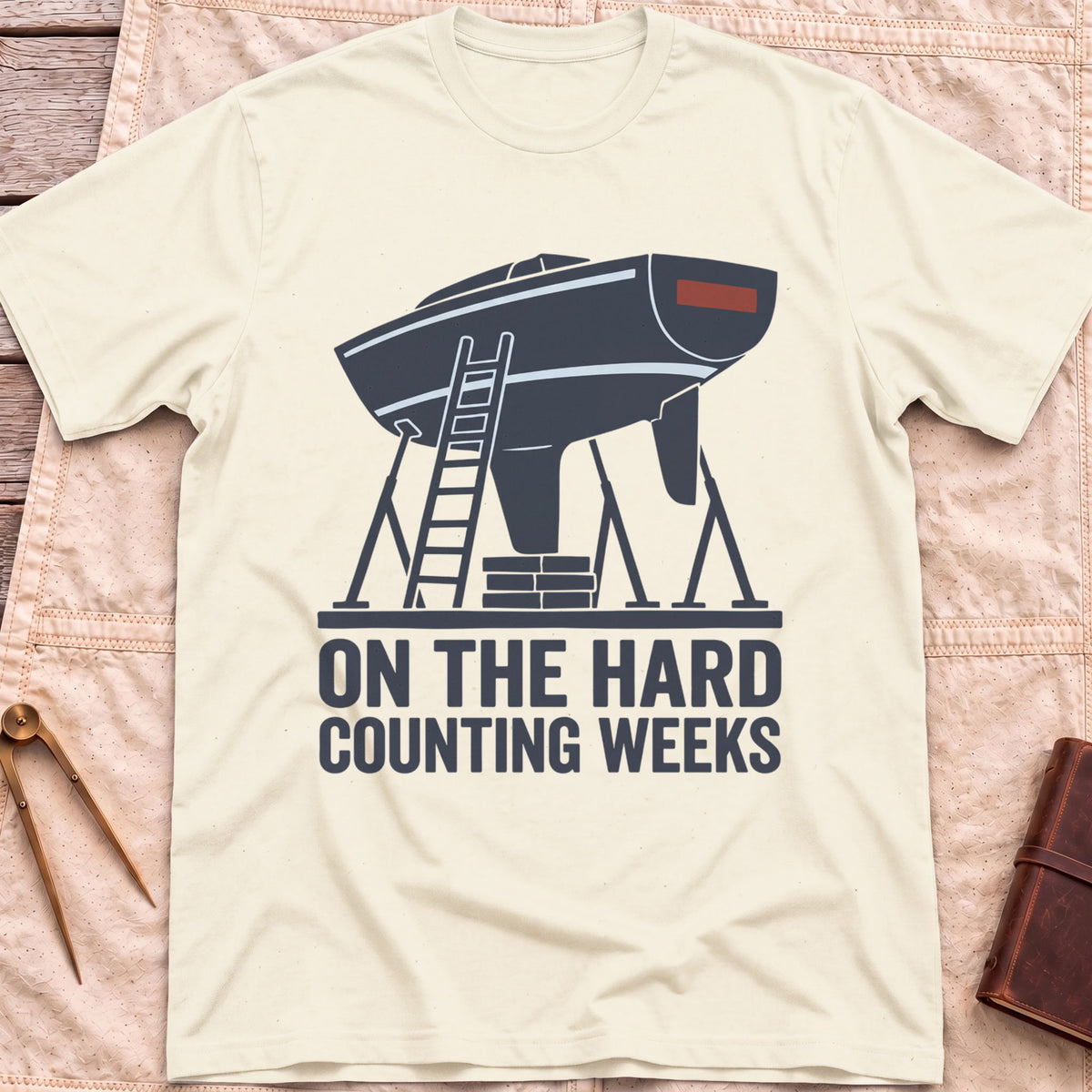 Front view of the natural On the Hard T-Shirt with a boat hull up on stands with a ladder against it, above the words Counting Weeks.