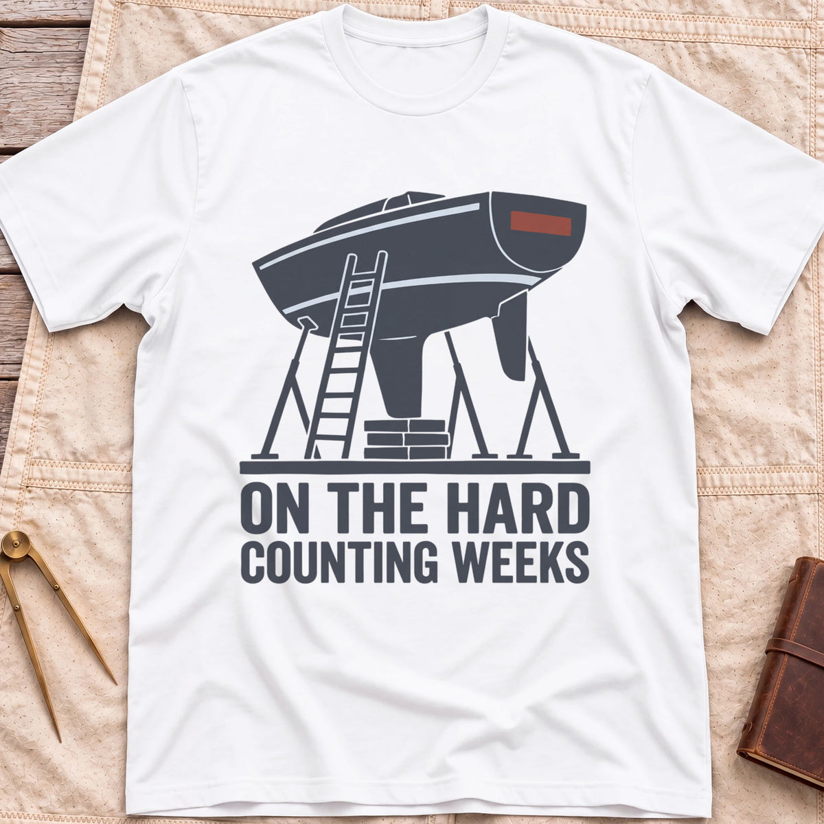 Front view of the white On the Hard T-Shirt with a boat hull up on stands with a ladder against it, above the words Counting Weeks.