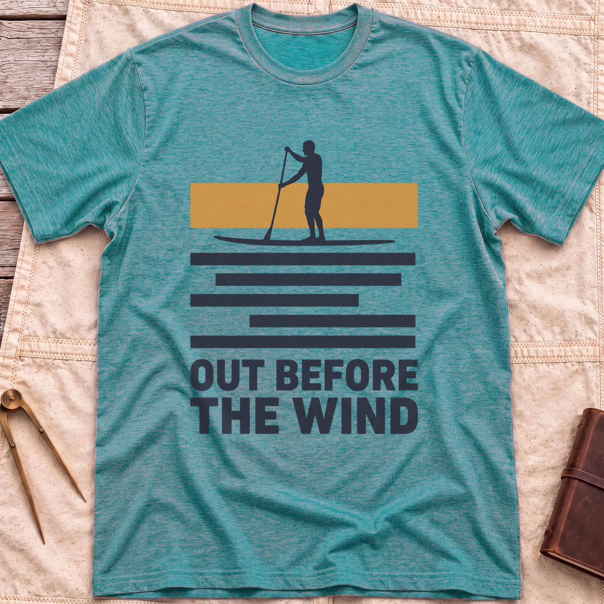 Front view of the heather galapagos blue Out Before the Wind T-Shirt with a paddleboarder silhouetted against a tan band above striped water and the words Out Before the Wind.