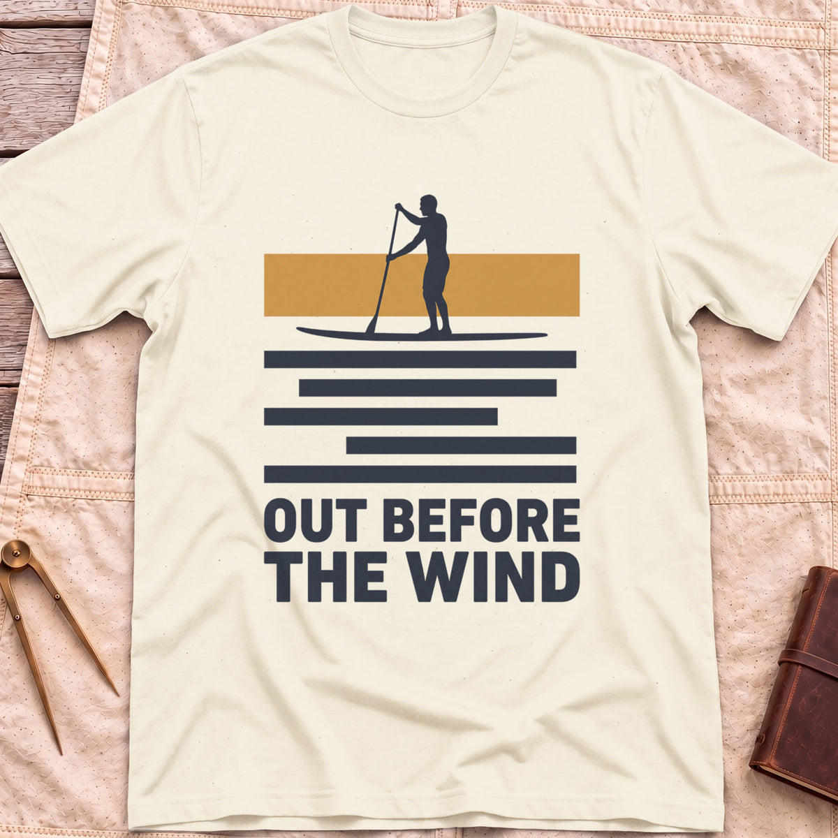 Front view of the natural Out Before the Wind T-Shirt with a paddleboarder silhouetted against a tan band above striped water and the words Out Before the Wind.