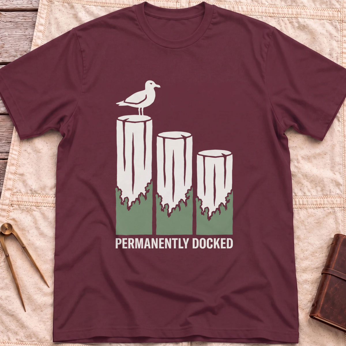 Front view of the maroon Permanently Docked T-Shirt with a seagull perched on three wooden pilings.