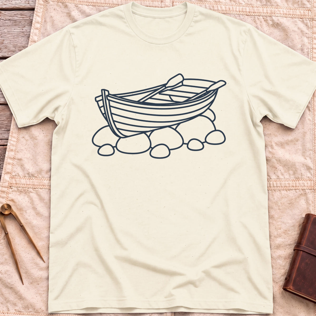 Front view of the natural Pulled Ashore T-Shirt with a line drawing of a rowing boat with oars, resting on rocks.