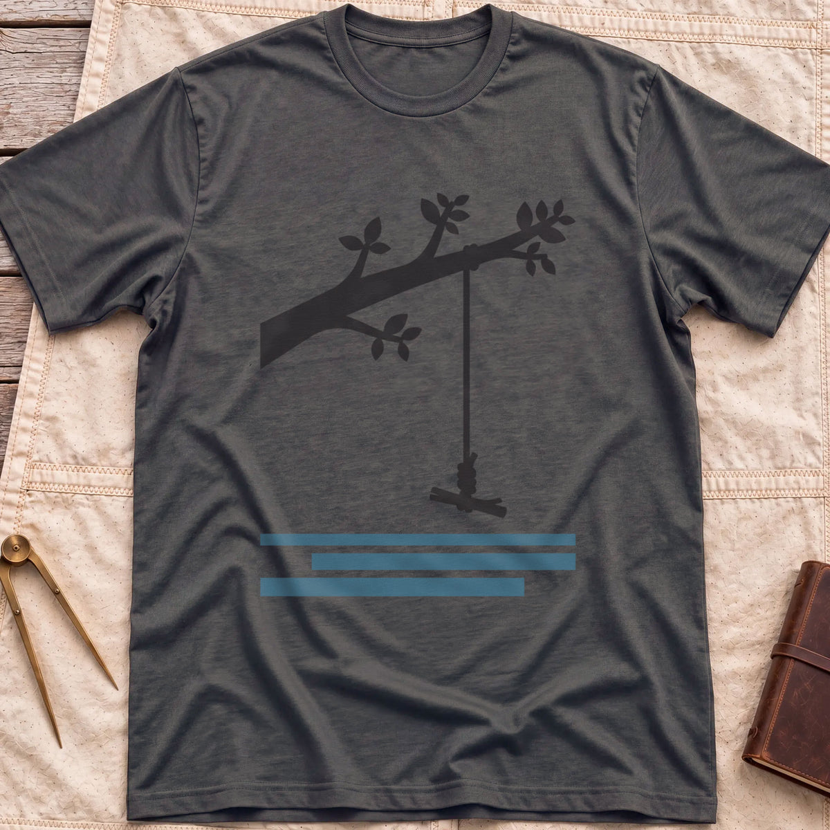 Front view of the dark heather Rope Swing T-Shirt with a rope swing hanging from a leafy branch above water lines.