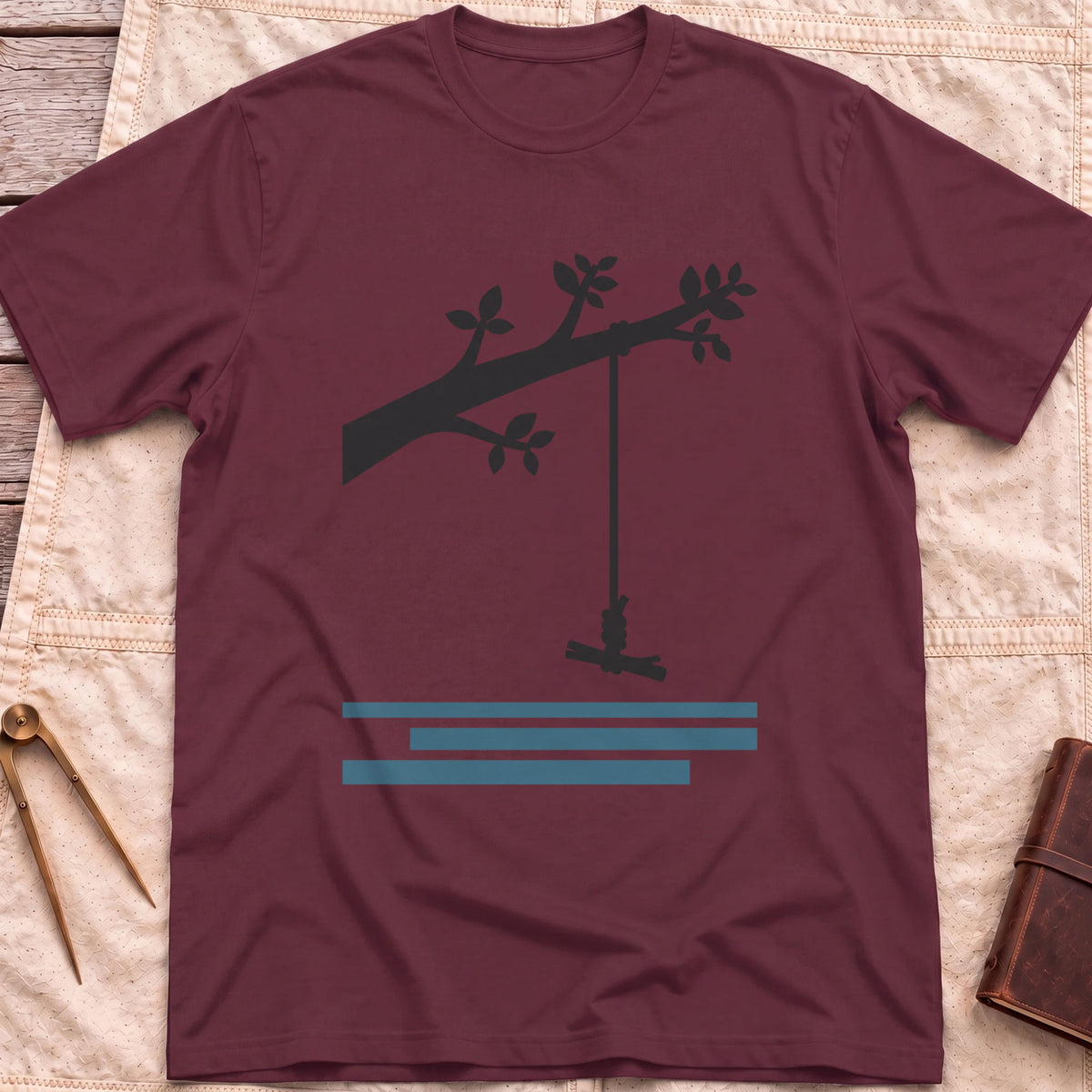 Front view of the maroon Rope Swing T-Shirt with a rope swing hanging from a leafy branch above water lines.
