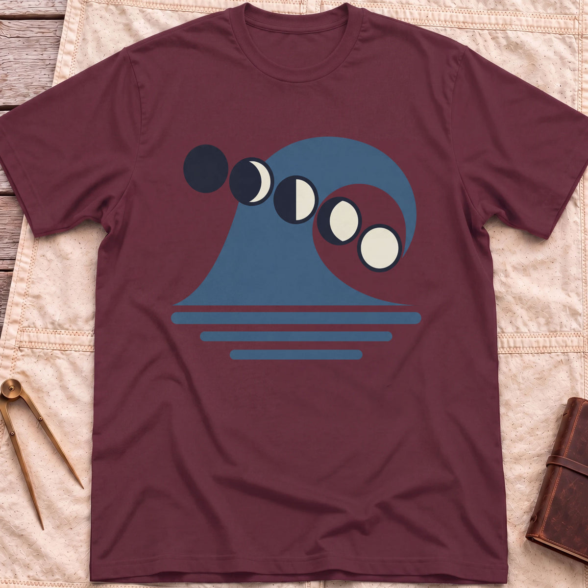 Front view of the maroon Ruled by the Tides T-Shirt with a stylised wave, a row of moon phases and horizontal water lines.