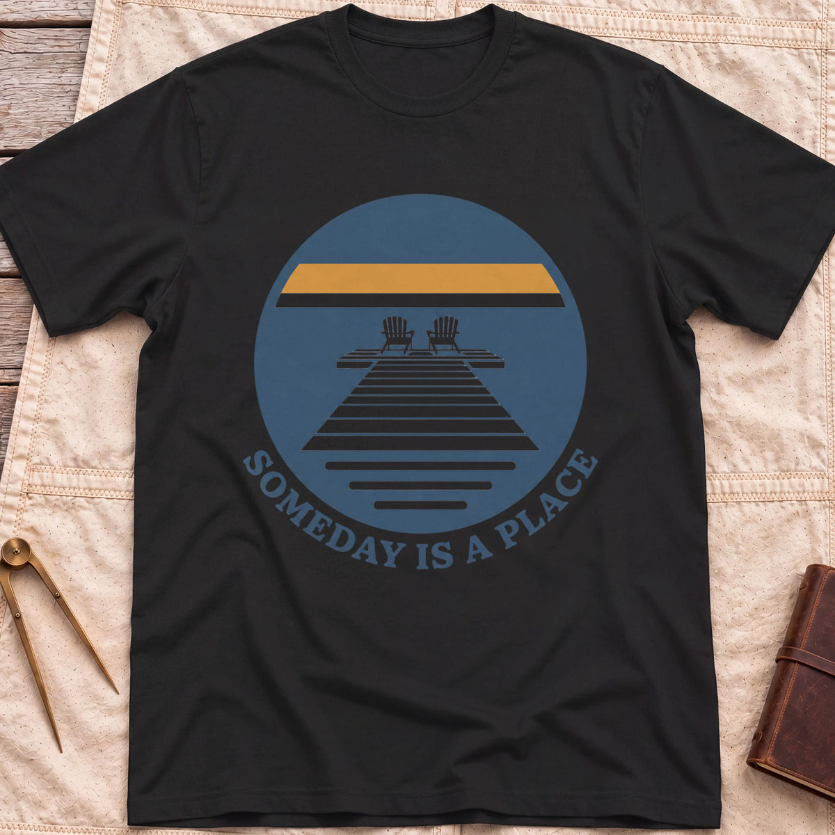 Front view of the black Someday Is a Place T-Shirt with a round badge showing two chairs at the end of a dock.