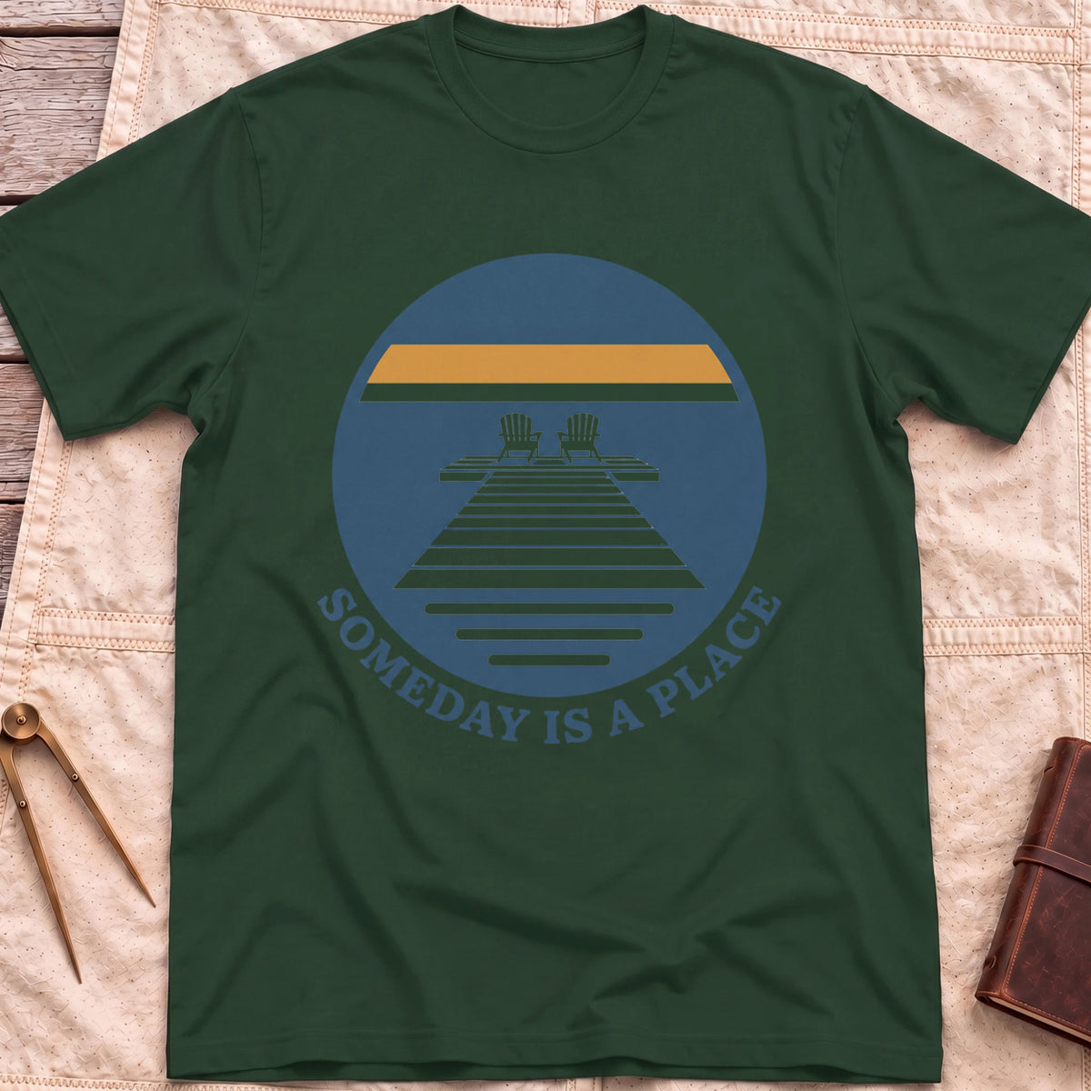 Front view of the forest green Someday Is a Place T-Shirt with a round badge showing two chairs at the end of a dock.