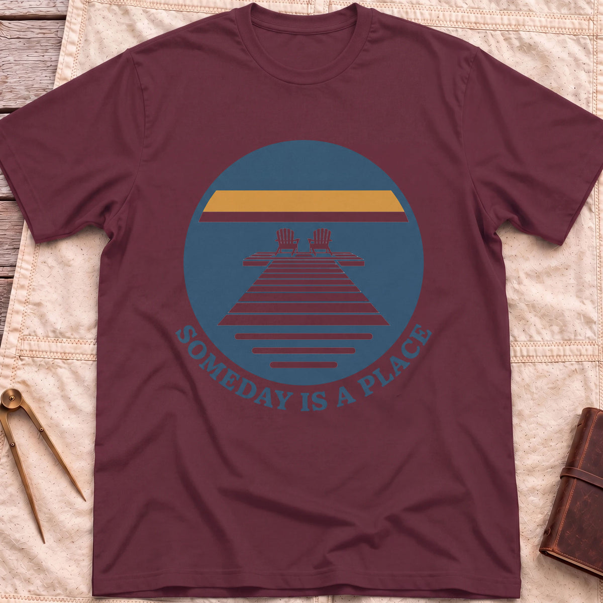 Front view of the maroon Someday Is a Place T-Shirt with a round badge showing two chairs at the end of a dock.
