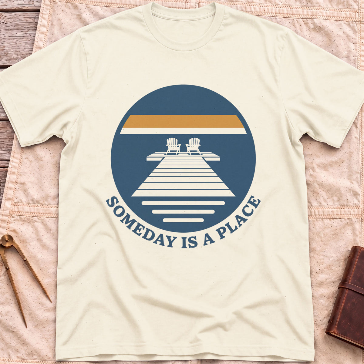 Front view of the natural Someday Is a Place T-Shirt with a round badge showing two chairs at the end of a dock.
