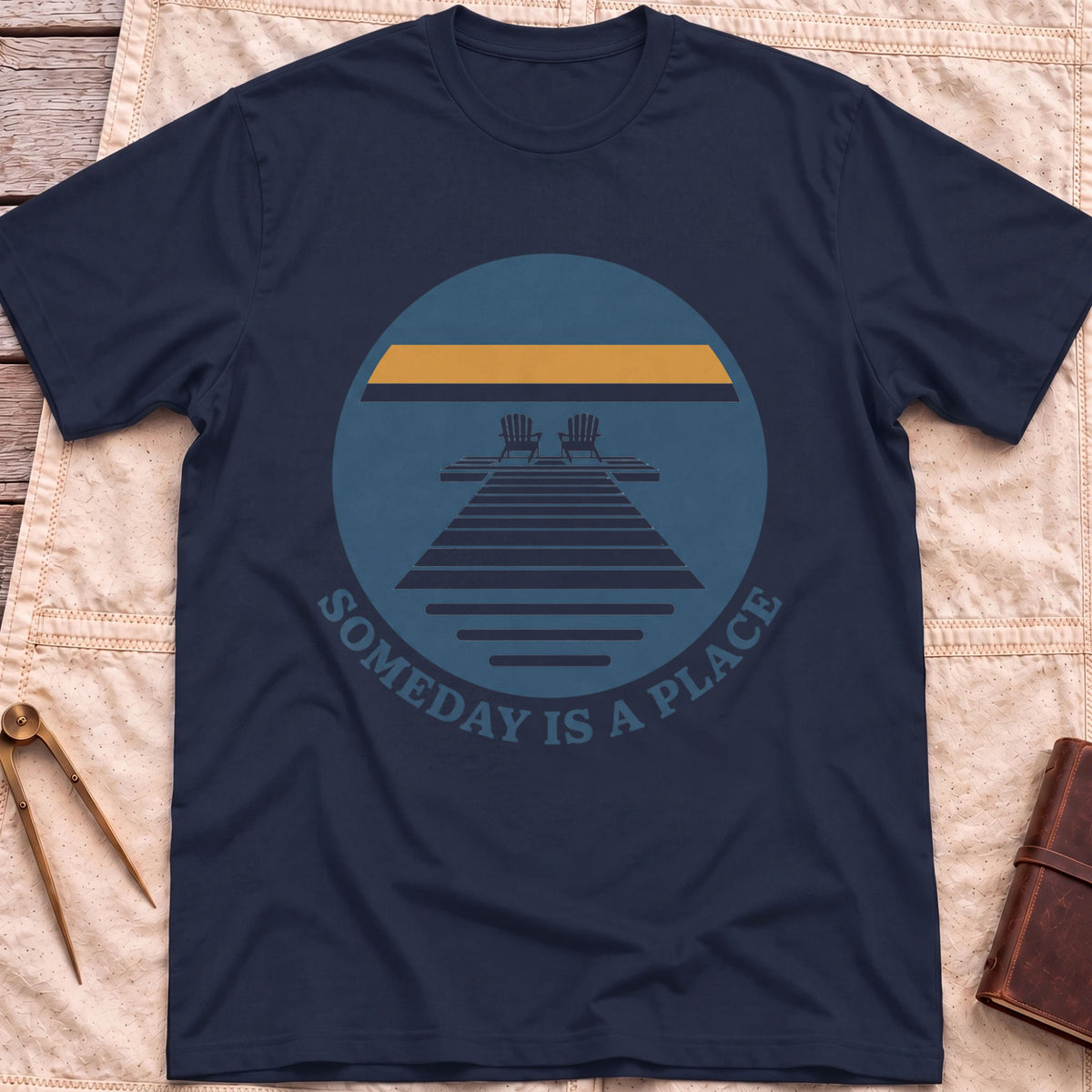 Front view of the navy Someday Is a Place T-Shirt with a round badge showing two chairs at the end of a dock.