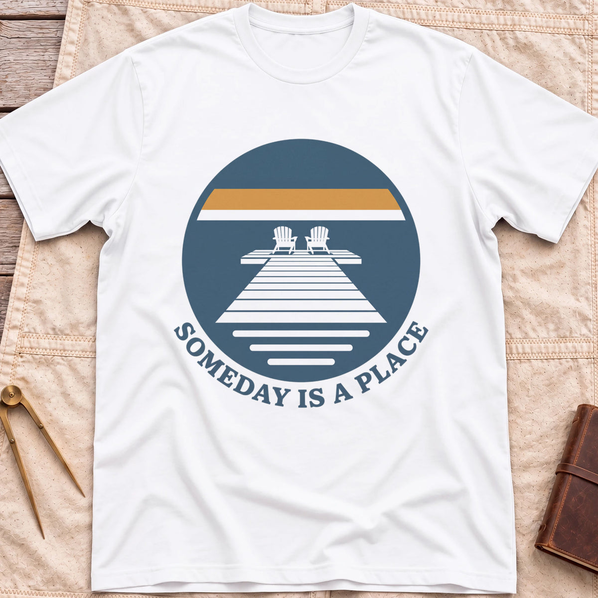 Front view of the white Someday Is a Place T-Shirt with a round badge showing two chairs at the end of a dock.