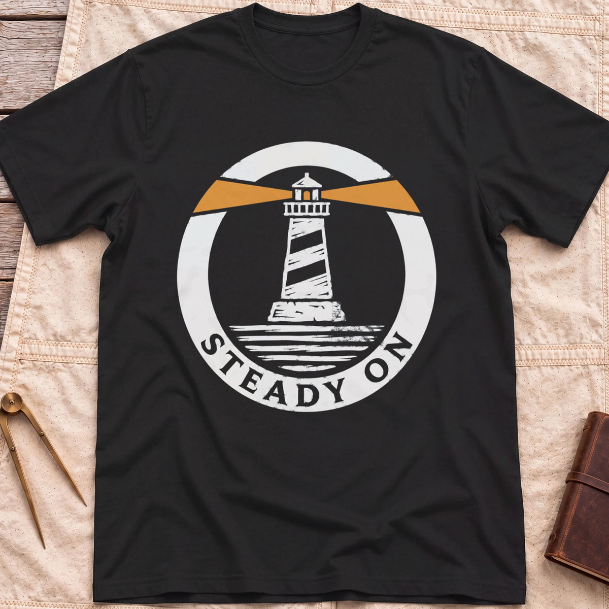 Front view of the black Steady On T-Shirt with a round badge showing a striped lighthouse and orange light beams over water.