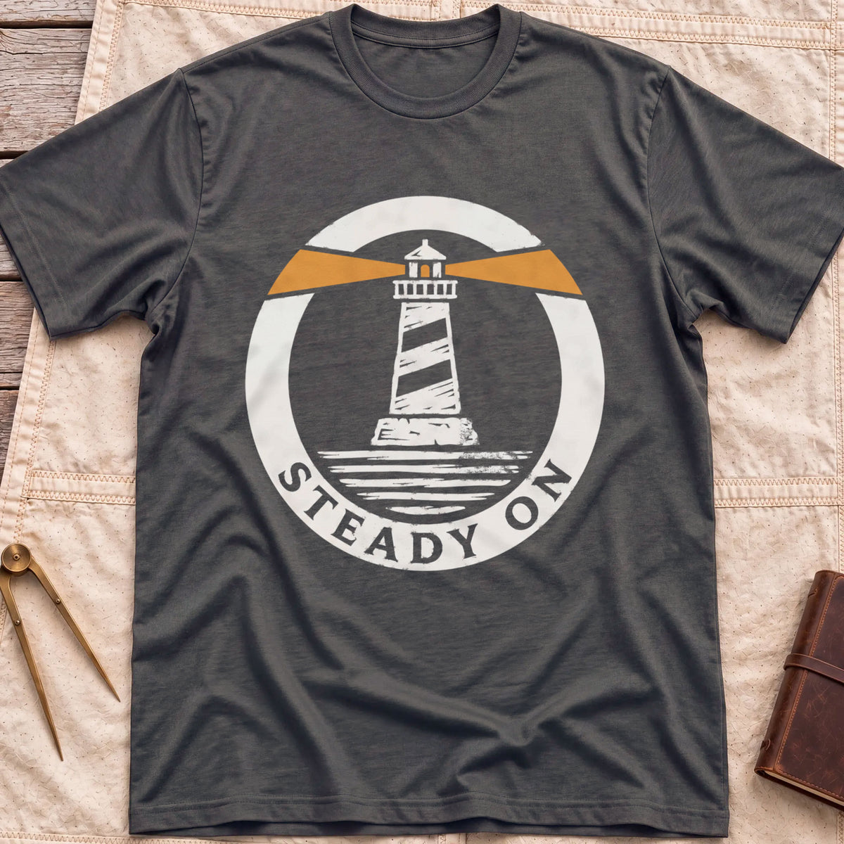 Front view of the dark heather Steady On T-Shirt with a round badge showing a striped lighthouse and orange light beams over water.