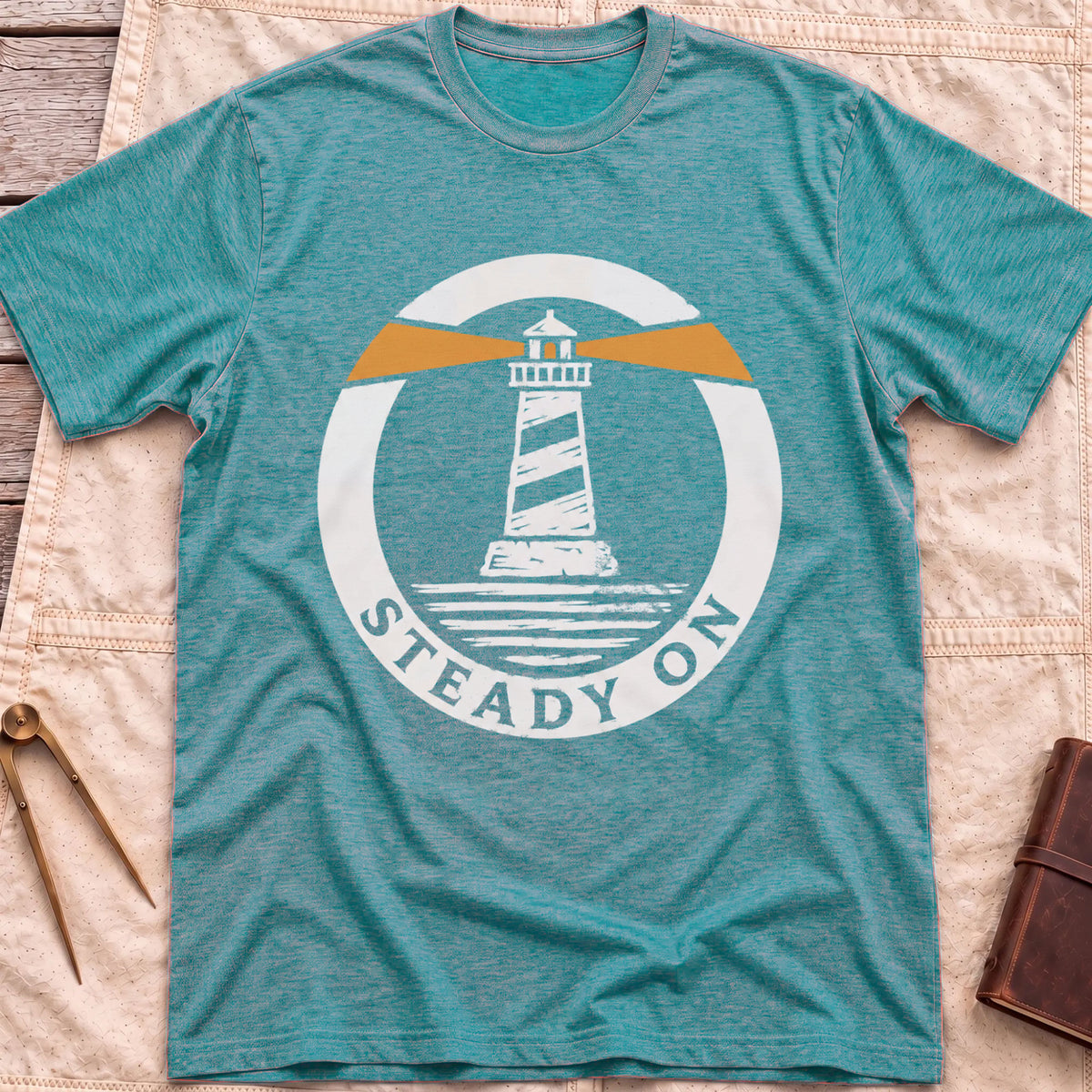 Front view of the heather galapagos blue Steady On T-Shirt with a round badge showing a striped lighthouse and orange light beams over water.
