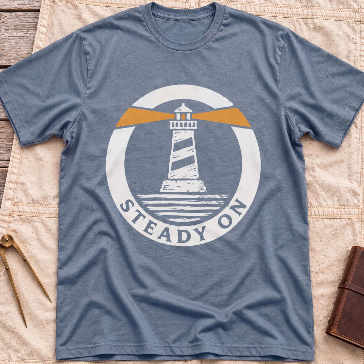 Front view of the heather indigo Steady On T-Shirt with a round badge showing a striped lighthouse and orange light beams over water.