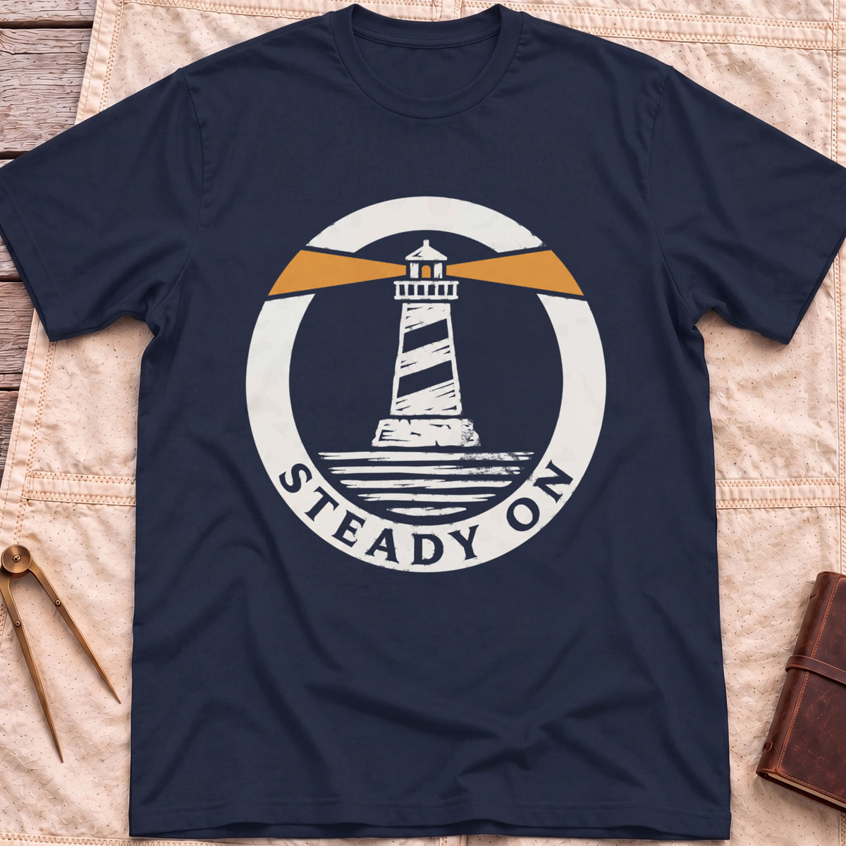 Front view of the navy Steady On T-Shirt with a round badge showing a striped lighthouse and orange light beams over water.