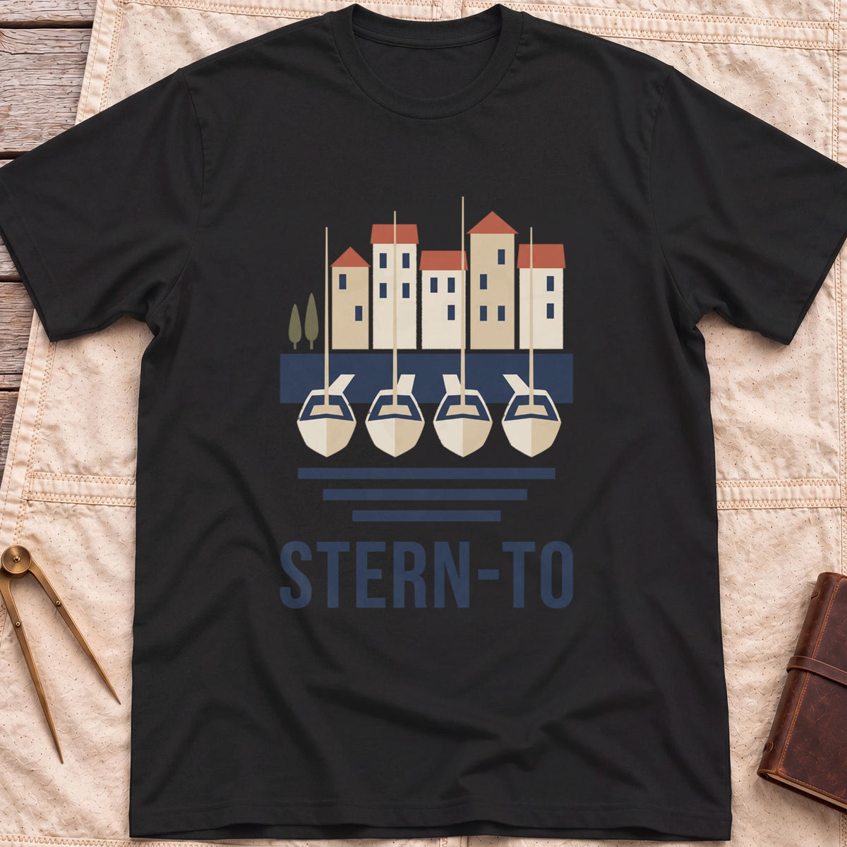 Front view of the black Stern-To T-Shirt with four boats moored stern-first to a quay below a row of harbour houses, above the word Stern-To.