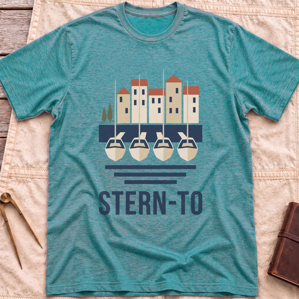 Front view of the heather galapagos blue Stern-To T-Shirt with four boats moored stern-first to a quay below a row of harbour houses, above the word Stern-To.
