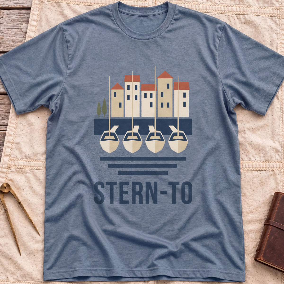 Front view of the heather indigo Stern-To T-Shirt with four boats moored stern-first to a quay below a row of harbour houses, above the word Stern-To.