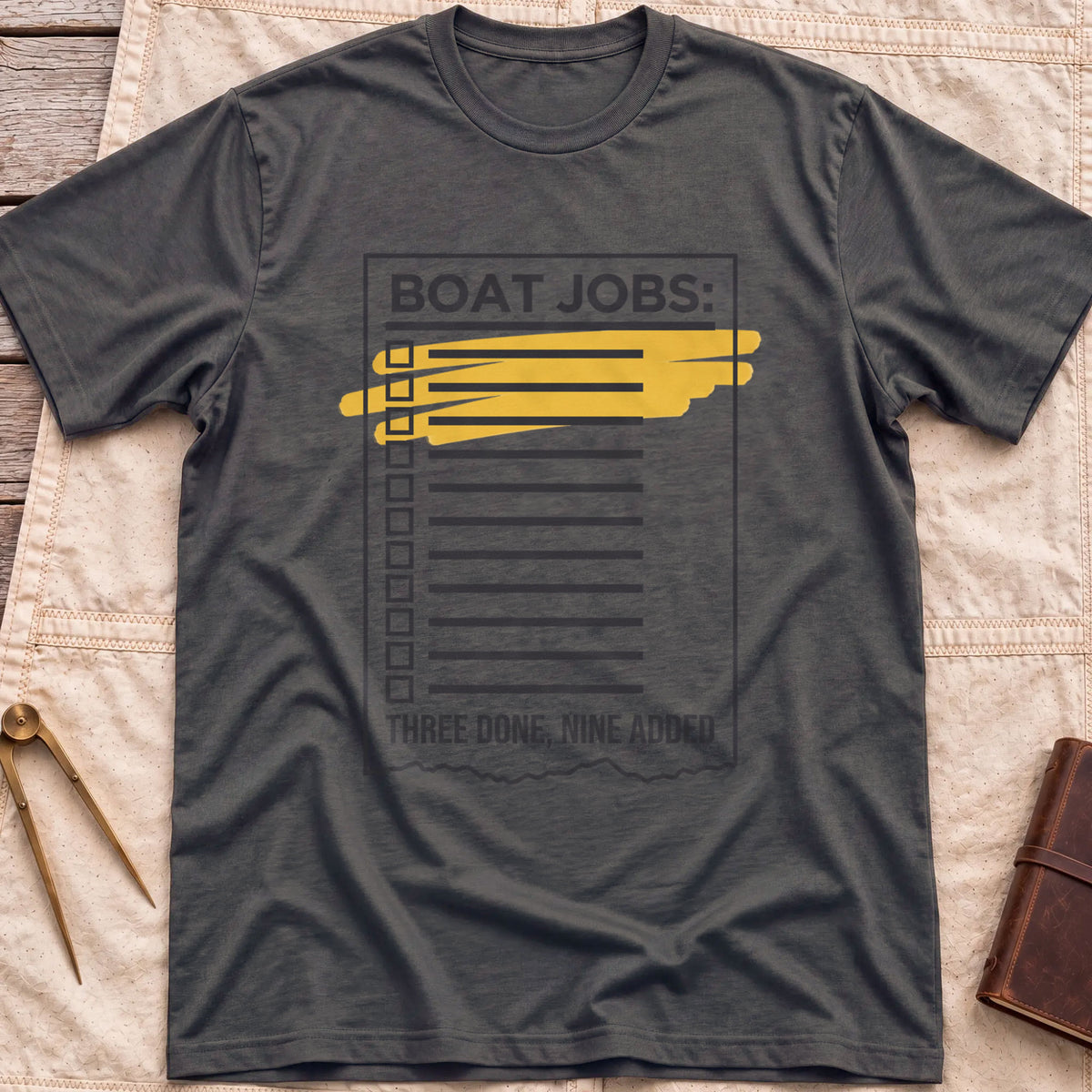 Front view of the dark heather Still on the List T-Shirt with a Boat Jobs checklist, its top items highlighted in yellow, above the words Three Done, Nine Added.