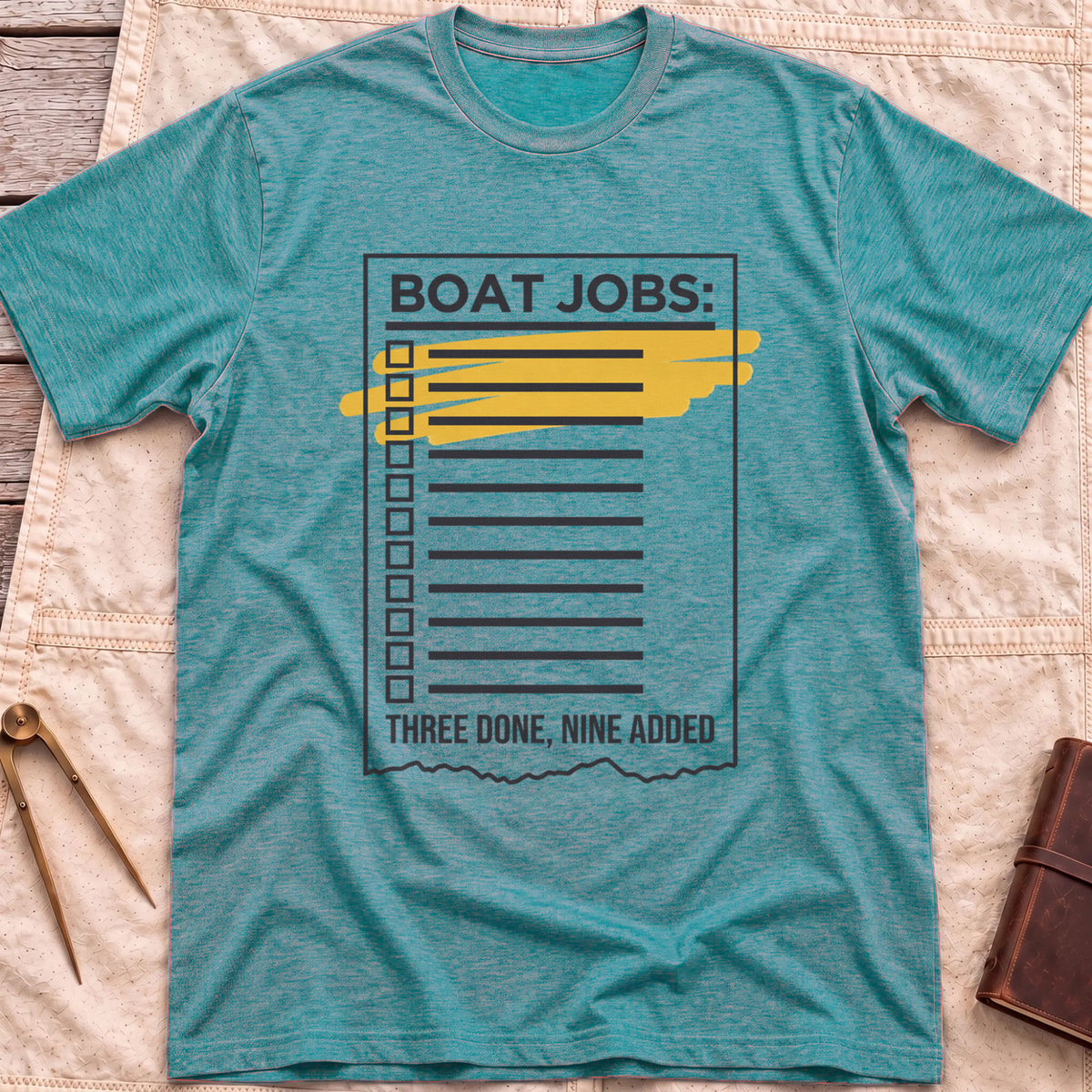 Front view of the heather galapagos blue Still on the List T-Shirt with a Boat Jobs checklist, its top items highlighted in yellow, above the words Three Done, Nine Added.