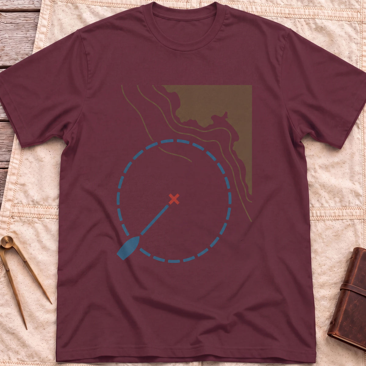 Front view of the maroon Swing Circle T-Shirt with a boat on a line inside a dashed circle around a red cross, beside a stretch of coastline.