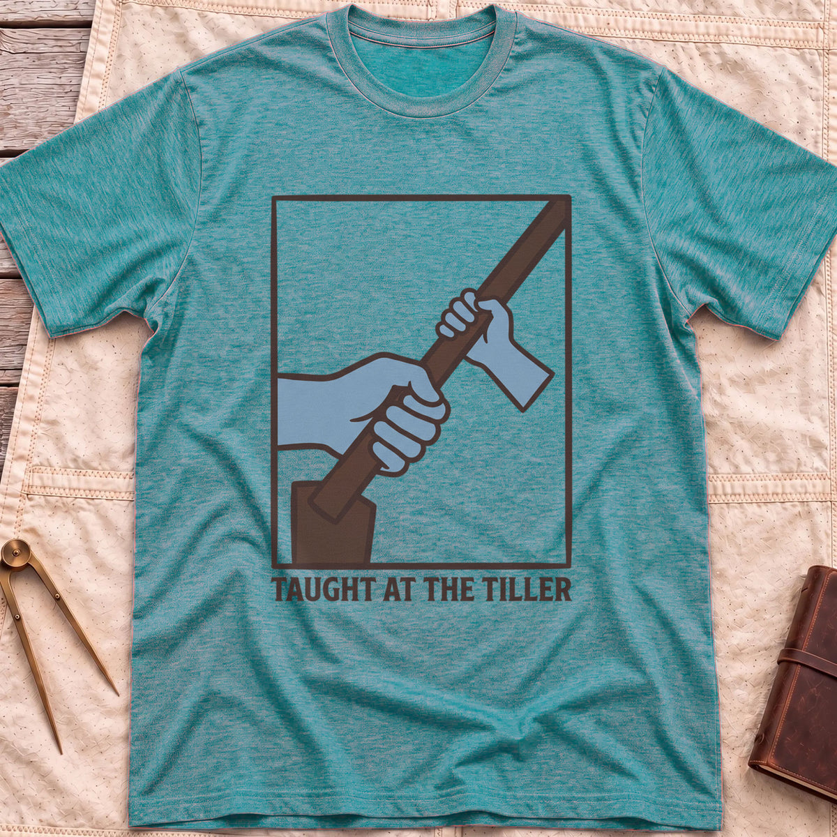 Front view of the heather galapagos blue Taught at the Tiller T-Shirt with a framed illustration of a larger hand and a smaller hand gripping a tiller together.