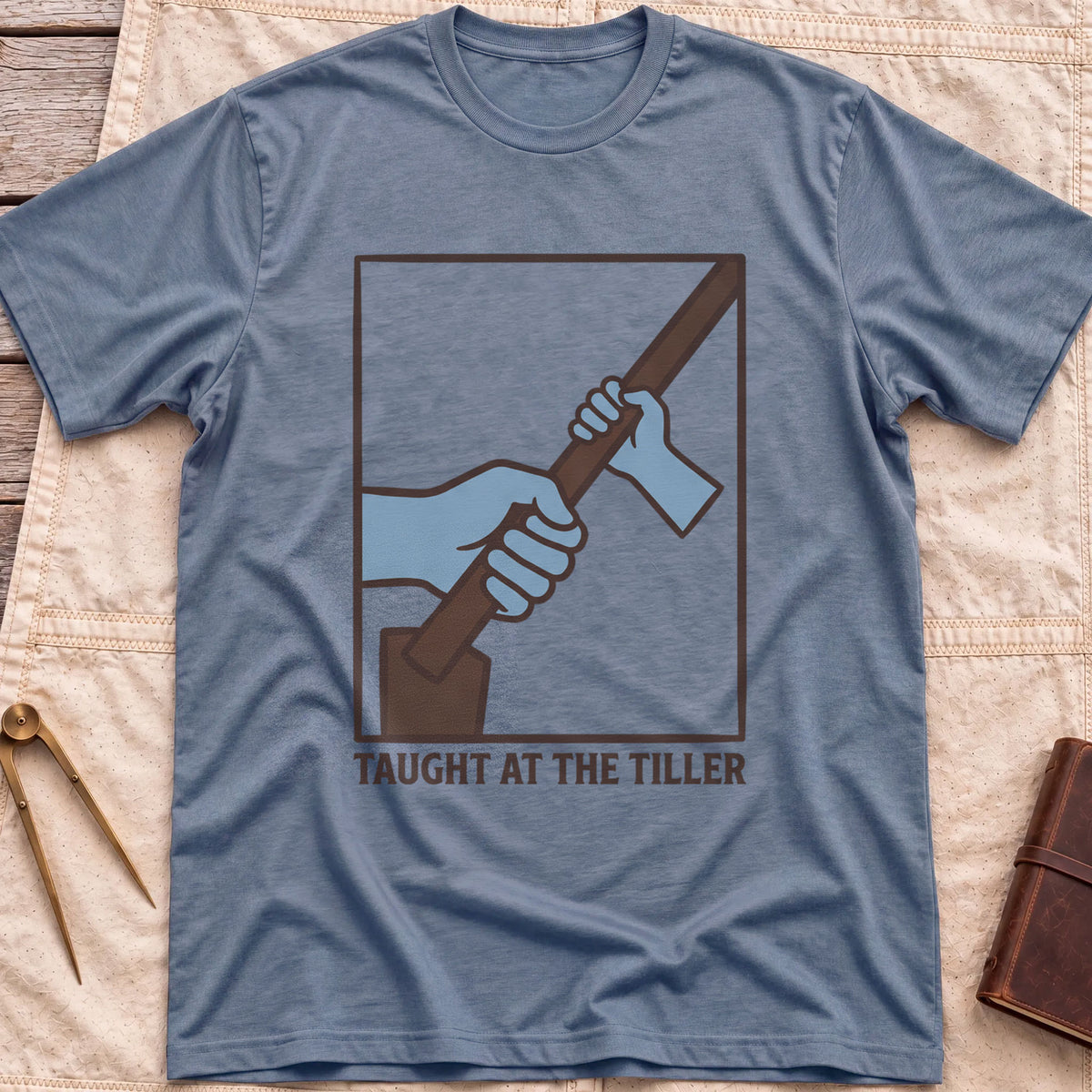 Front view of the heather indigo Taught at the Tiller T-Shirt with a framed illustration of a larger hand and a smaller hand gripping a tiller together.