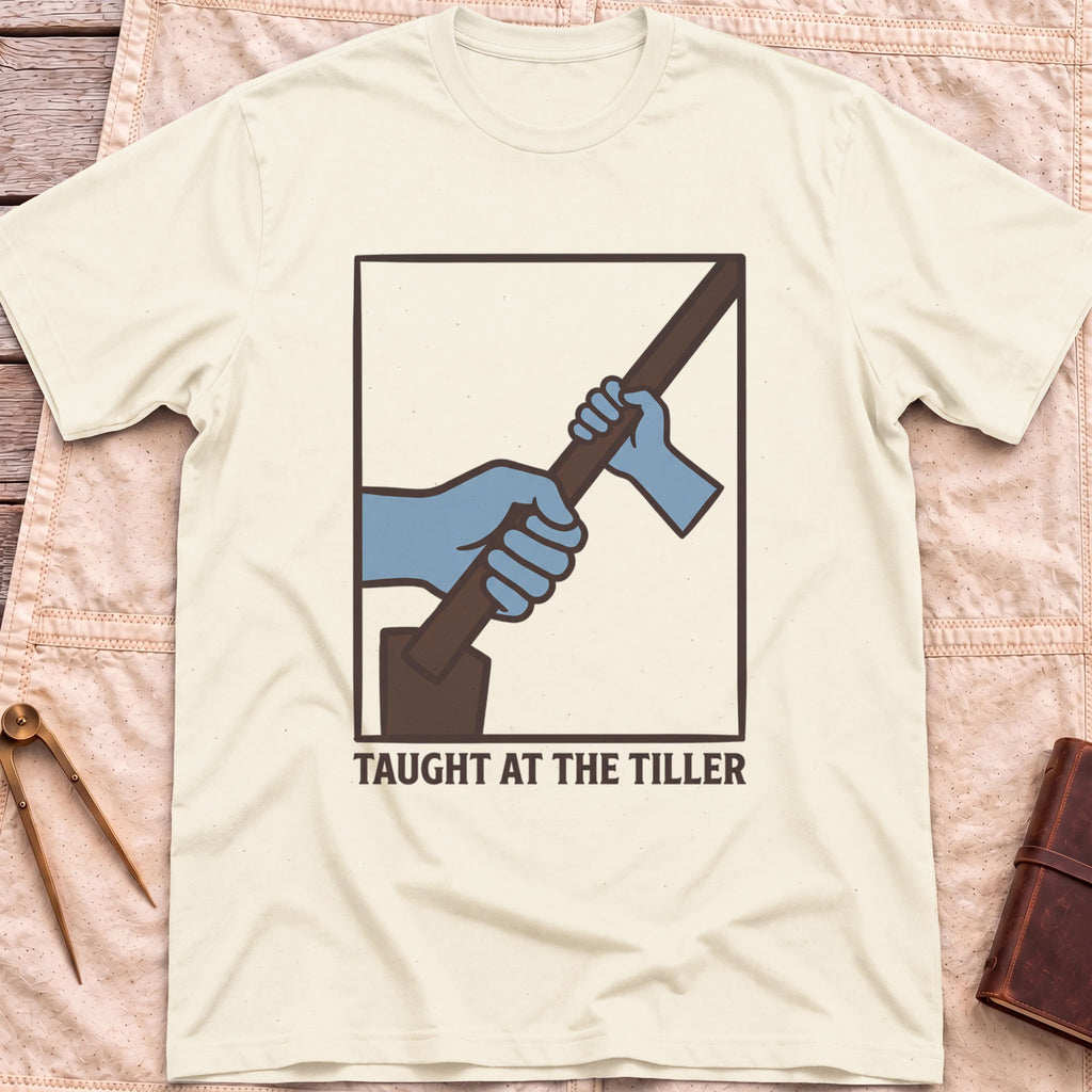 Front view of the natural Taught at the Tiller T-Shirt with a framed illustration of a larger hand and a smaller hand gripping a tiller together.