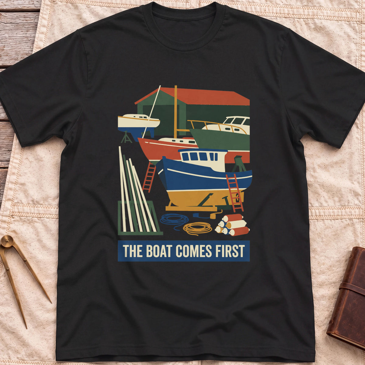 Front view of the black The Boat Comes First T-Shirt with a colourful boatyard scene of boats on stands, ladders, timber and coiled rope.