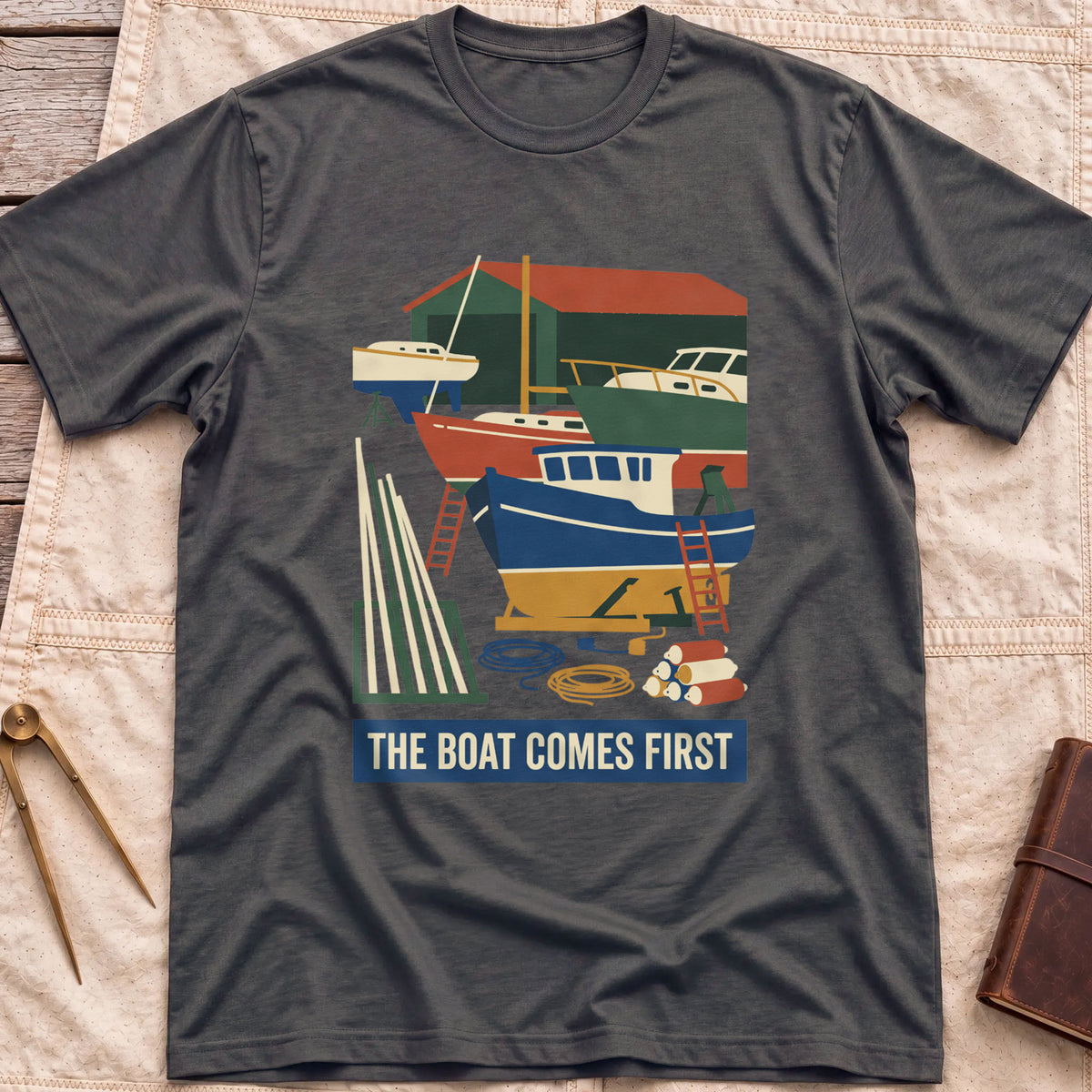 Front view of the dark heather The Boat Comes First T-Shirt with a colourful boatyard scene of boats on stands, ladders, timber and coiled rope.