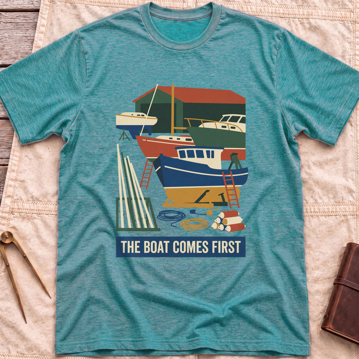 Front view of the heather galapagos blue The Boat Comes First T-Shirt with a colourful boatyard scene of boats on stands, ladders, timber and coiled rope.